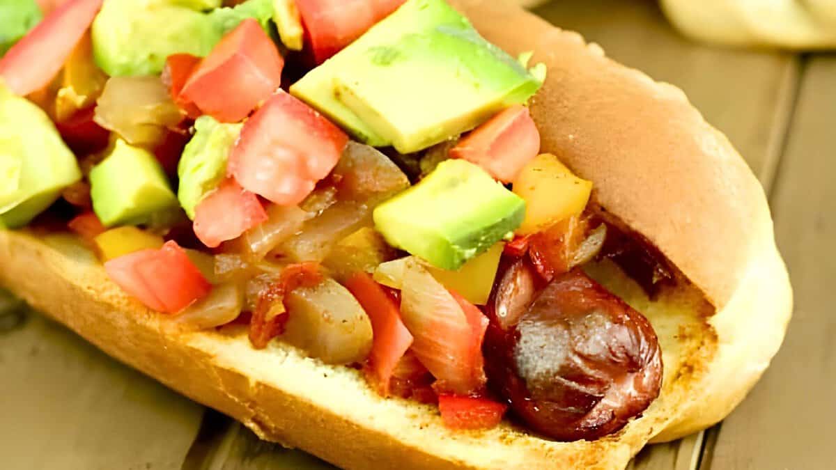 A hot dog in a bun is topped with chopped onions, tomatoes, and avocado.