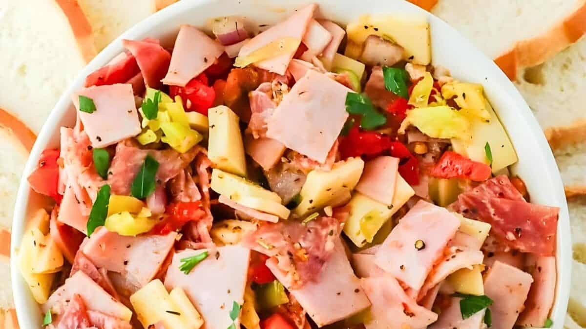 Close-up of an antipasto salad with slices of ham, cheese, tomato, onions, peperoncini, and garnished with parsley, served in a white bowl.