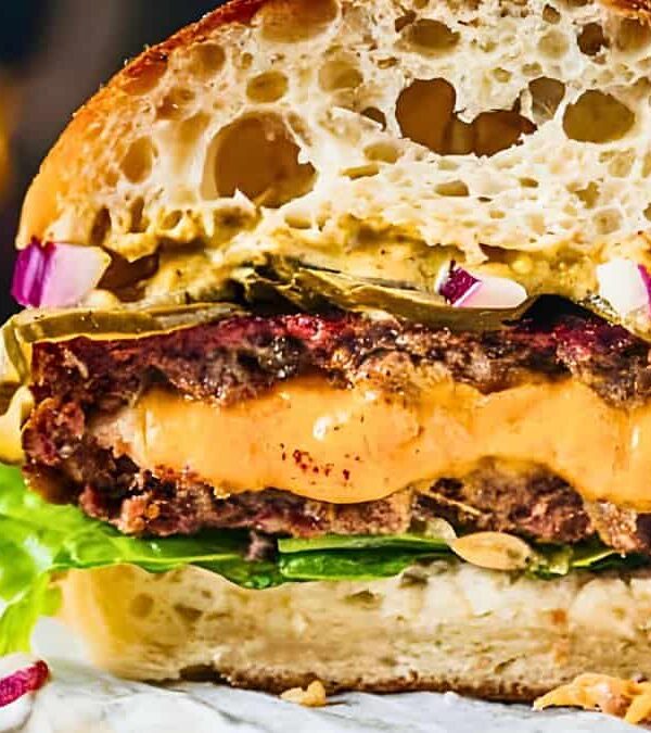 Close-up of a cut burger showcasing layers of lettuce, a juicy beef patty with melted cheese, pickles, sliced red onions, and sauce on a toasted bun, with blurred background elements.