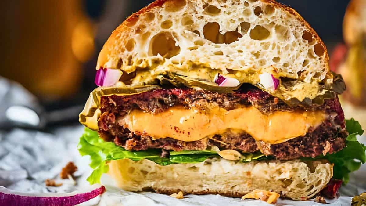 Close-up of a cut burger showcasing layers of lettuce, a juicy beef patty with melted cheese, pickles, sliced red onions, and sauce on a toasted bun, with blurred background elements.