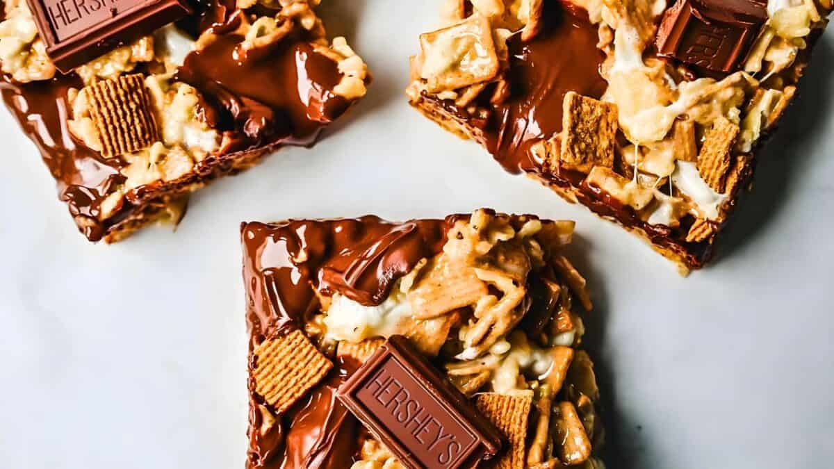 Three chocolate-covered cereal and marshmallow squares topped with small chocolate pieces, arranged on a white surface.