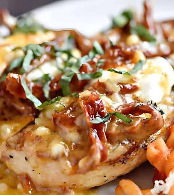 A close-up of a plate of grilled chicken topped with melted cheese, caramelized onions, and fresh herbs, accompanied by seasoned curly fries.