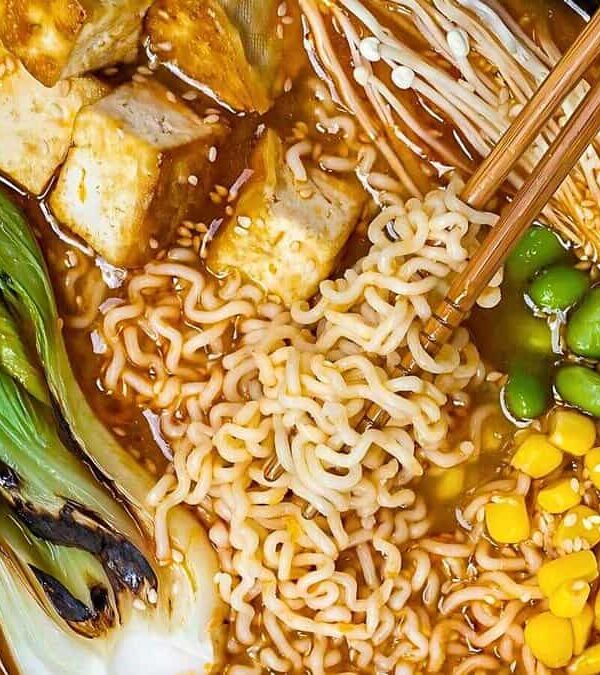 A bowl of ramen with noodles, tofu, bok choy, edamame, corn, and mushrooms. Chopsticks rest on the bowl.
