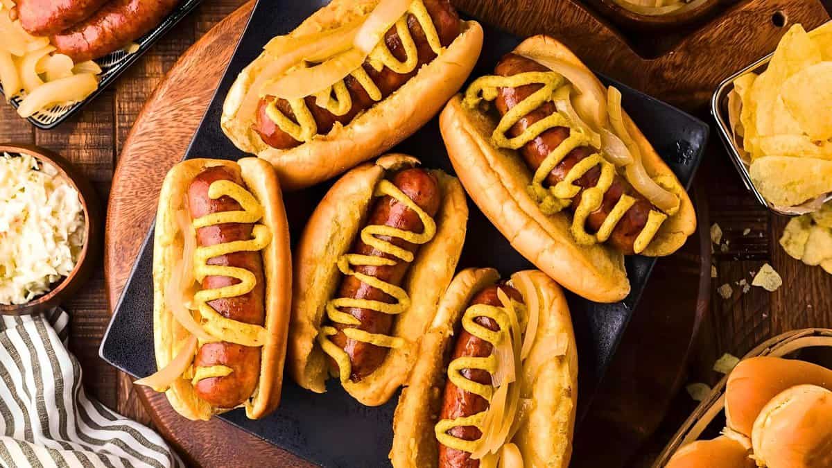 Five hot dogs with mustard and onions on a black platter, surrounded by bowls of coleslaw and potato chips on a wooden table.