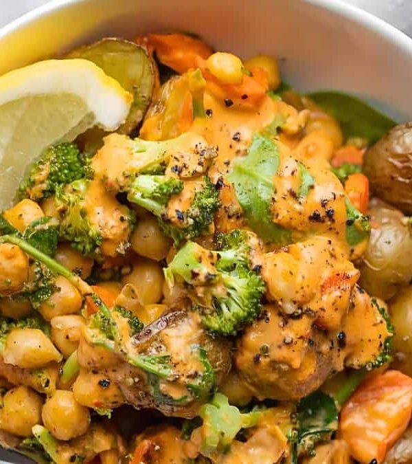 A white bowl filled with a mixed vegetable and chickpea salad, garnished with herbs and a lemon wedge, covered in a creamy orange sauce.