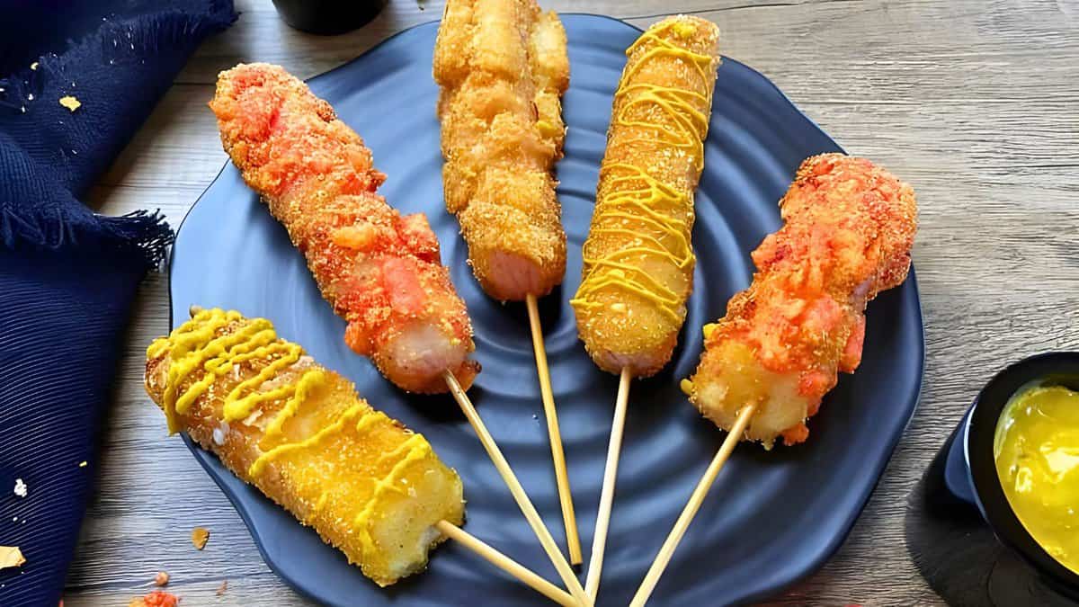 A black plate holds five corn dogs, each coated in different toppings and drizzled with mustard. A small sauce dish and a dark cloth napkin are placed beside the plate on a wooden table.