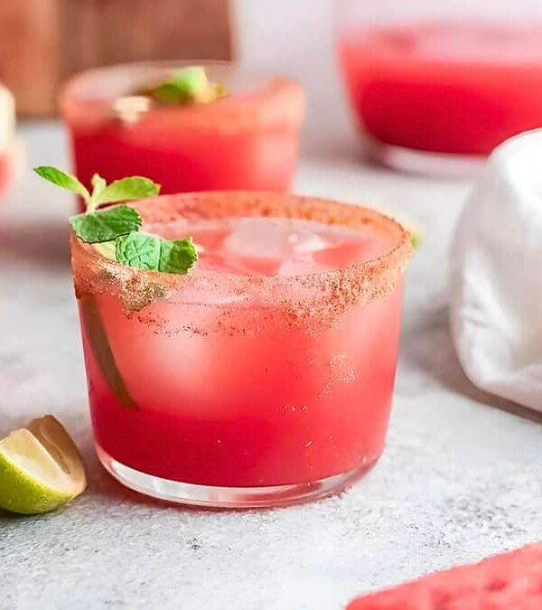 Three glasses of watermelon juice garnished with mint leaves, with sliced lime and a watermelon in the background on a light surface.