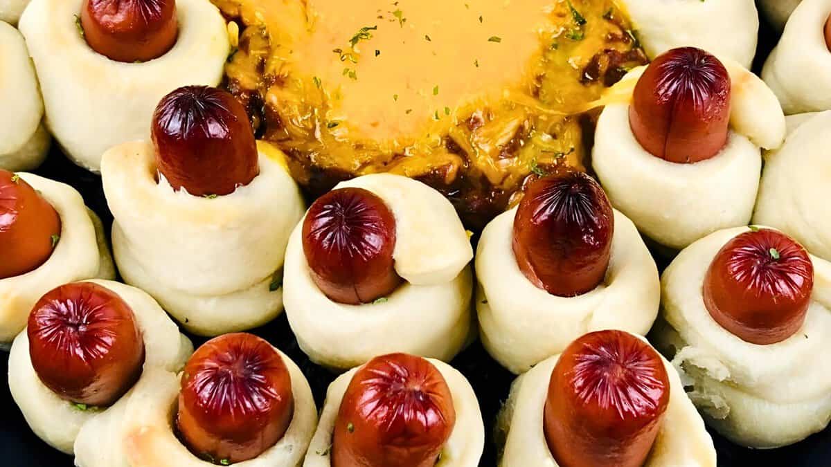 A close-up of pigs in a blanket arranged in a circular shape with cheese dip in the center. The dish consists of sausages wrapped in dough, baked until golden brown.