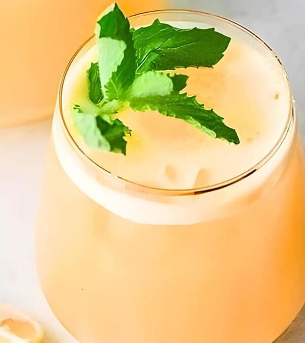 A frothy orange drink garnished with a sprig of mint is served in a clear glass, placed on a light-colored surface next to a sliced lemon and a wooden straw.