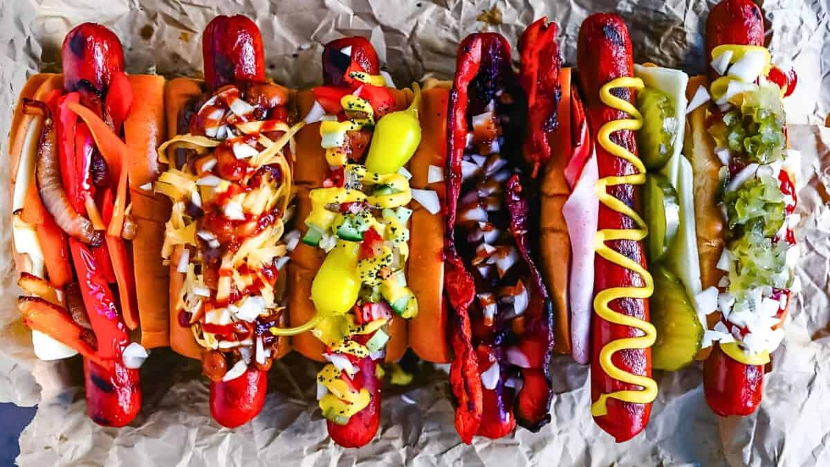 A row of seven hot dogs on parchment paper, each topped with a variety of condiments such as onions, peppers, mustard, relish, and a pepperoncini.