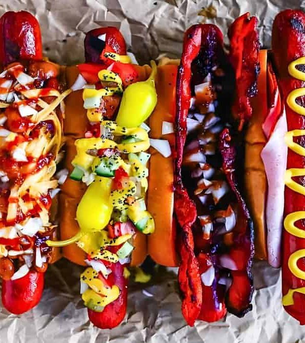 A row of seven hot dogs on parchment paper, each topped with a variety of condiments such as onions, peppers, mustard, relish, and a pepperoncini.