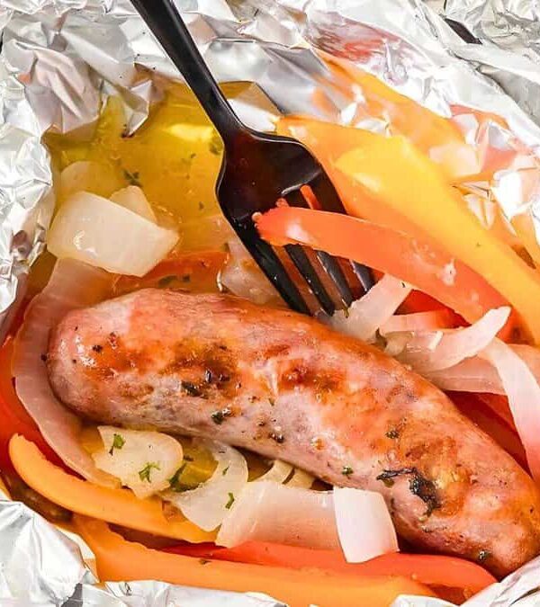 Grilled sausage with sliced bell peppers and onions in aluminum foil, with a black plastic fork.