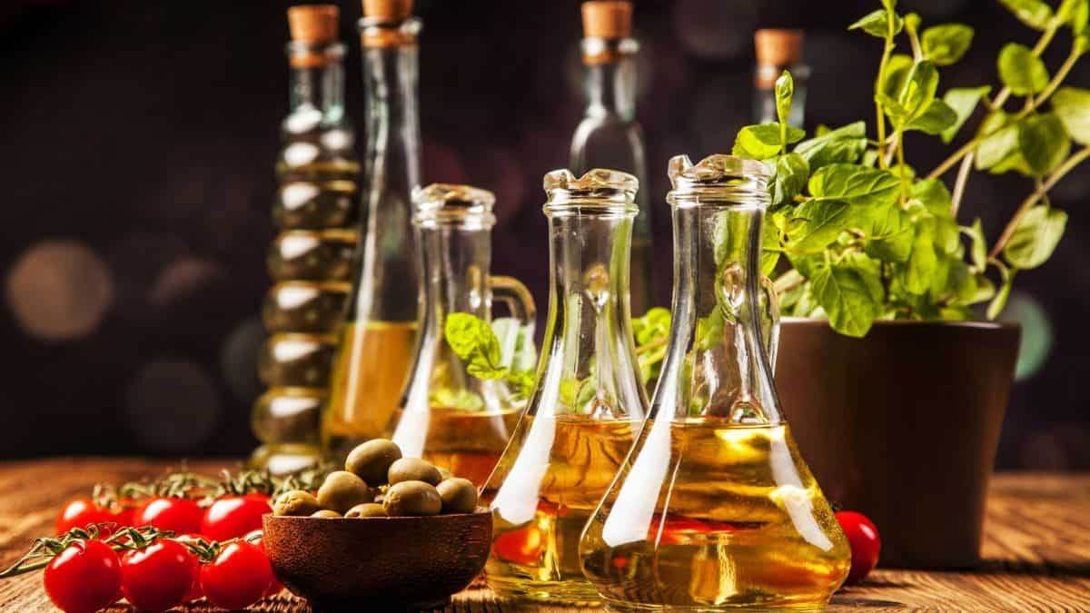 Bottles of olive oil, tomatoes, a bowl of olives, and fresh herbs in pots on a wooden surface with a blurred background.