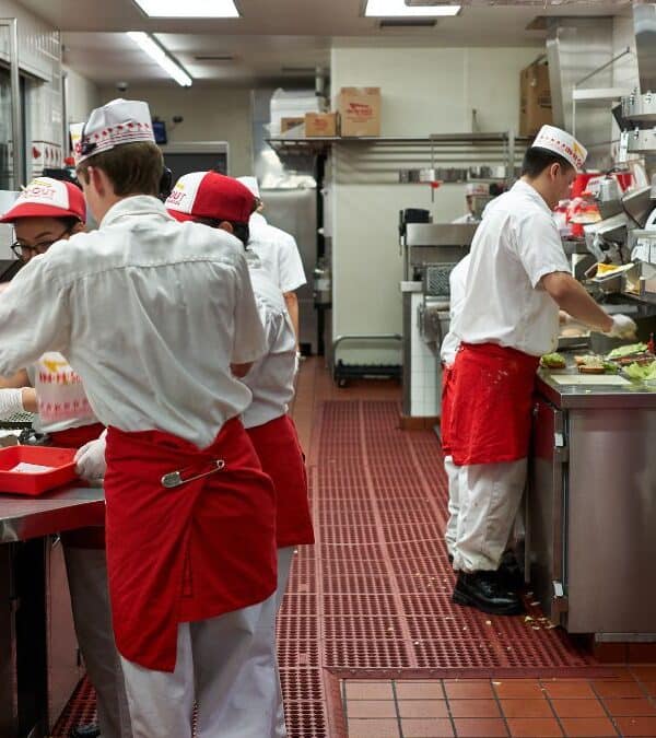 Employees in red aprons and white uniforms working in a fast-food kitchen, preparing and assembling meals on a tiled floor with various kitchen equipment and ingredients around.