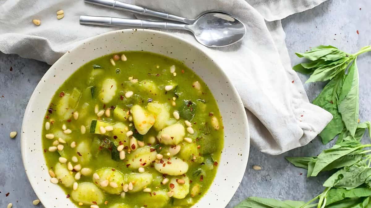 A bowl of green vegetable soup garnished with pine nuts and red pepper flakes, accompanied by a spoon, set on a light blue textured surface.