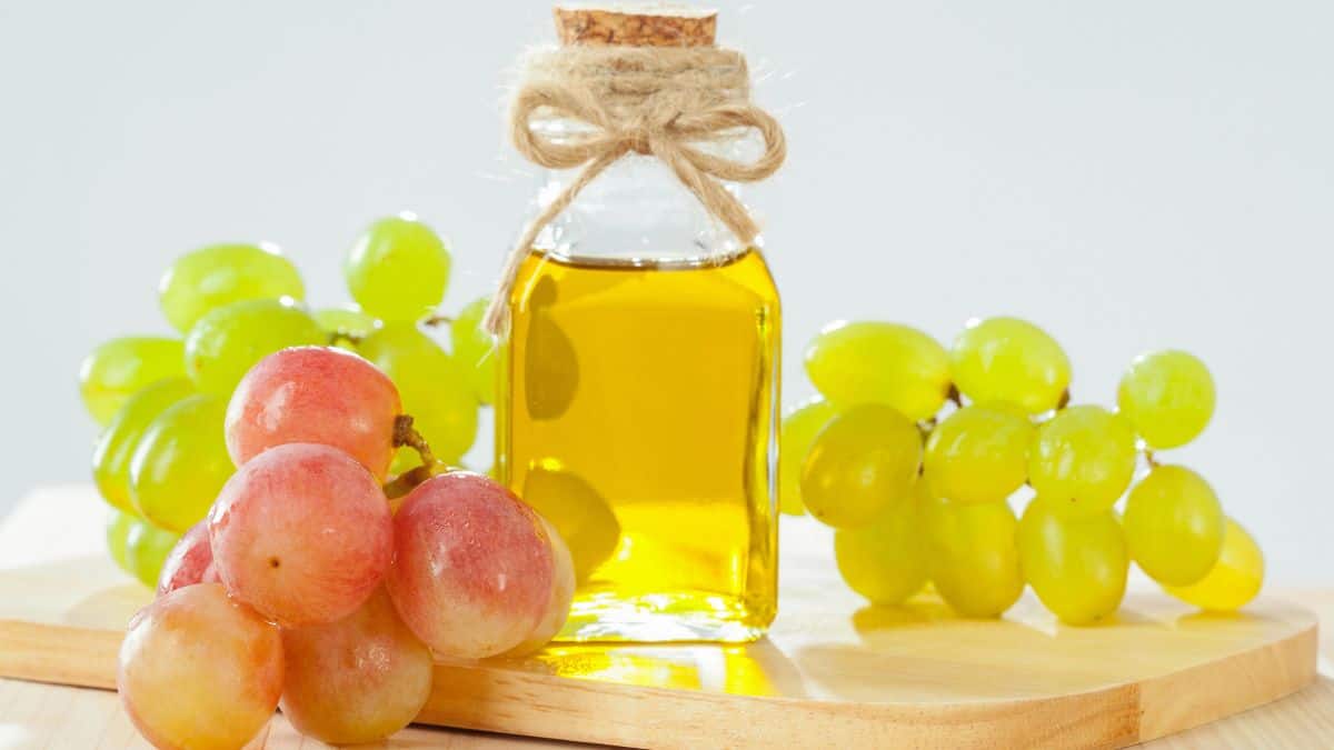 A bottle of grape seed oil with a cork stopper, tied with a piece of twine, is placed on a wooden board alongside bunches of green and red grapes.
