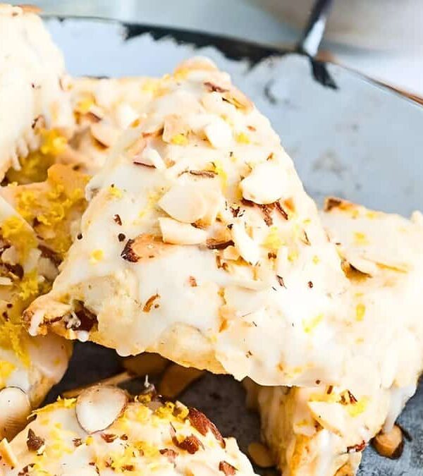 Sliced almond-topped lemon scones on a glass plate, with visible zest and a drizzle of glaze, emphasizing a fresh, homemade appearance.