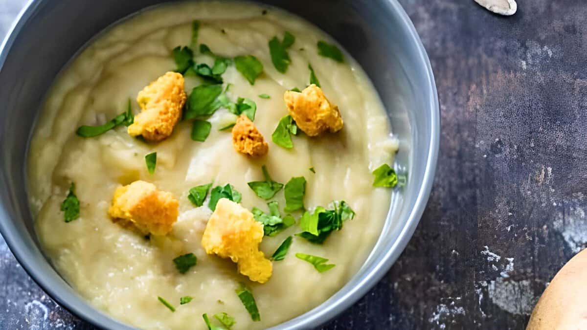 A bowl of creamy soup garnished with croutons and chopped herbs, presented on a rustic dark surface.