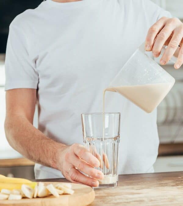 A person is pouring a light-colored beverage from a measuring cup into a glass. A cutting board with sliced bananas is on the counter in front of them.
