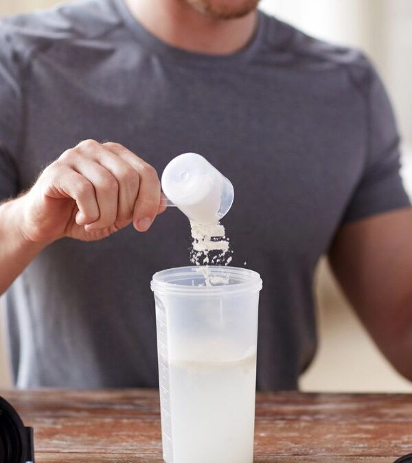 A person in a gray shirt is scooping protein powder from a large container into a shaker bottle filled with liquid, while seated at a table.