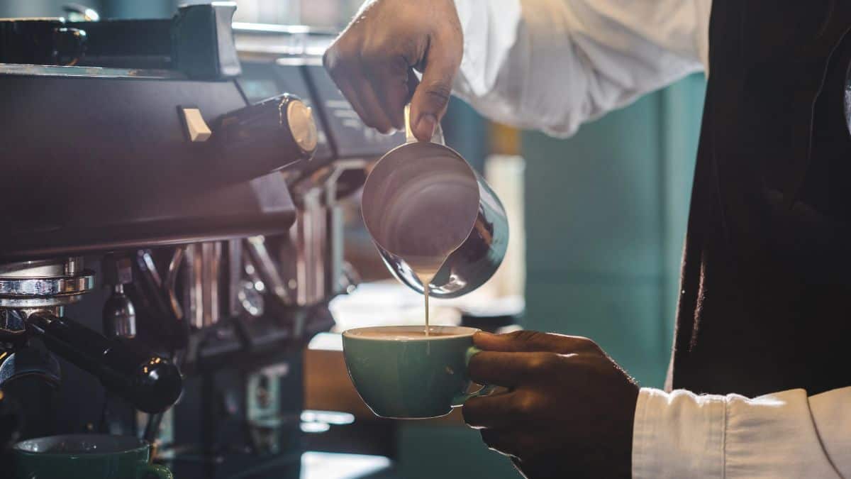 A barista pours milk into a coffee cup from a jug to create latte art, positioned beside an espresso machine.