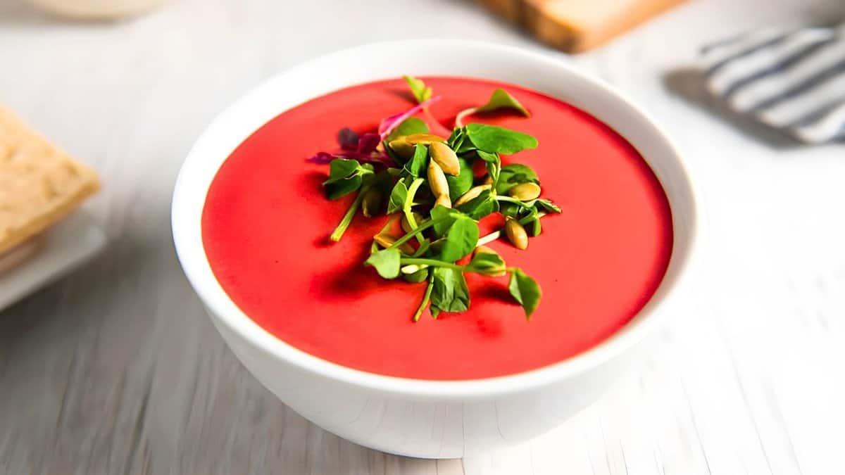 A bowl of vibrant red beet soup garnished with greens and nuts, served on a white table with a napkin and bread in the background.