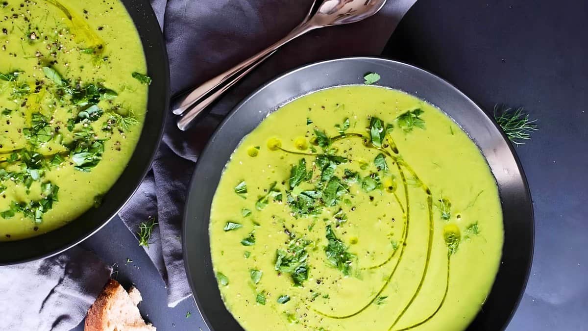 Two bowls of vibrant green pea soup garnished with herbs, served with a spoon on a dark tablecloth.