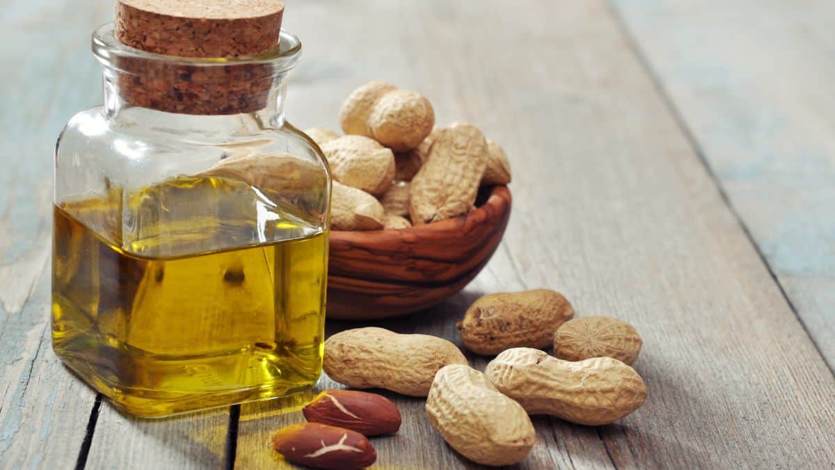 A glass bottle of peanut oil with a cork stopper stands next to a wooden bowl filled with peanuts and several loose peanuts on a rustic wooden surface.