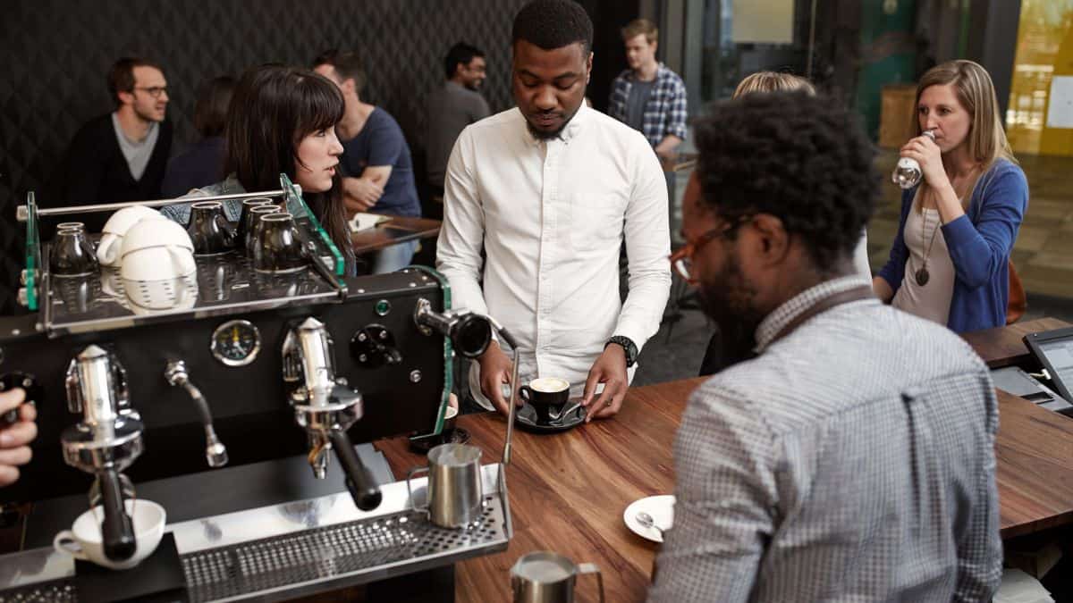 A diverse group of people interacting at a coffee shop with one man preparing coffee at an espresso machine.