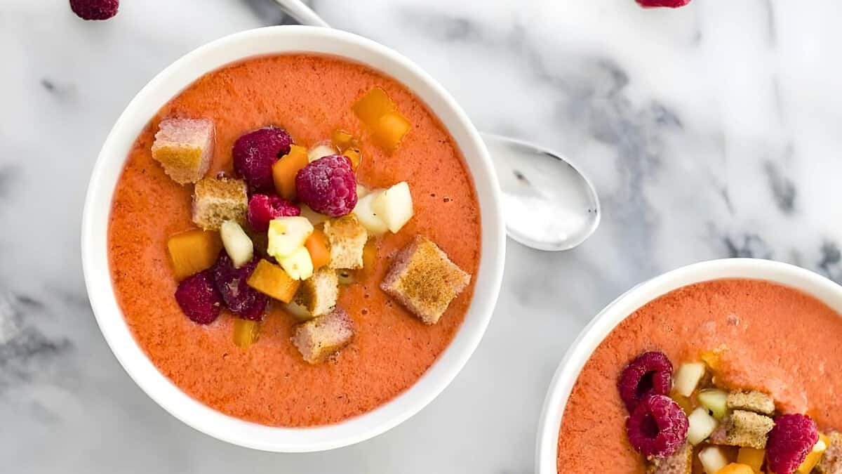 Two bowls of fruit gazpacho topped with diced mango, raspberries, and croutons on a marble surface.