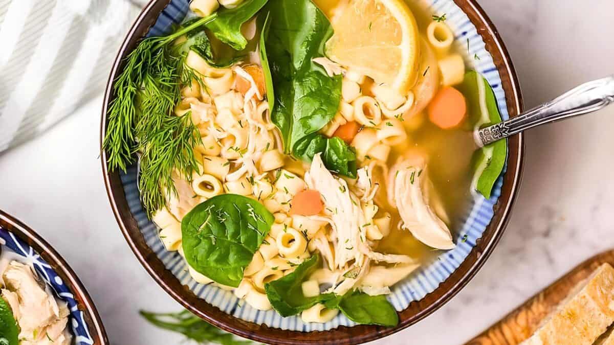 A bowl of chicken noodle soup with spinach, carrots, and herbs, served with a slice of lemon, viewed from above.