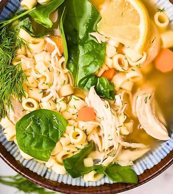 A bowl of chicken noodle soup with spinach, carrots, and herbs, served with a slice of lemon, viewed from above.