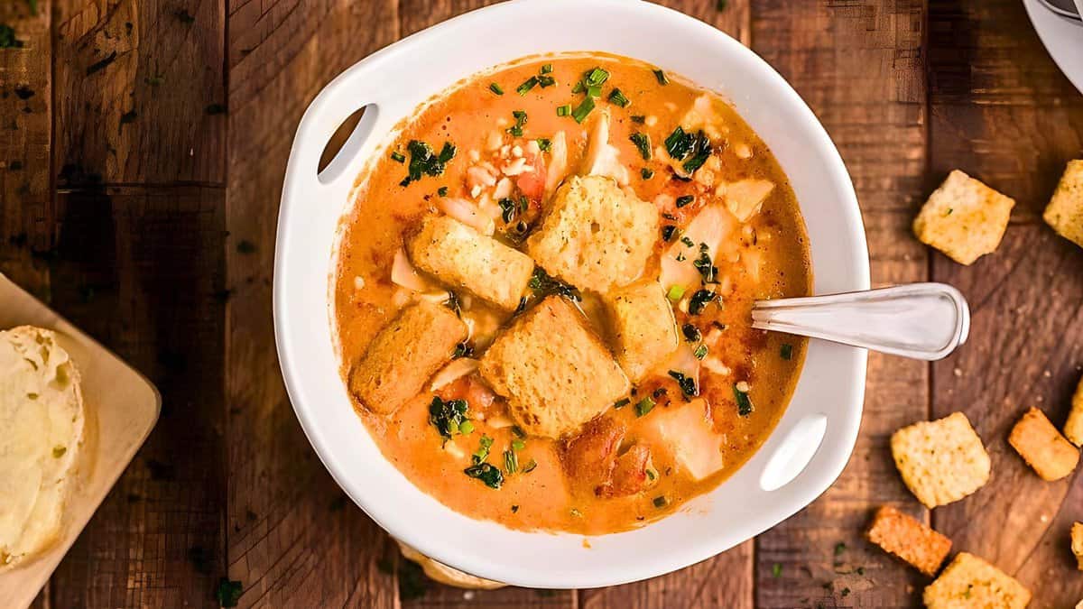 A bowl of tomato soup garnished with herbs and croutons, served on a wooden table with extra croutons and bread on the side.