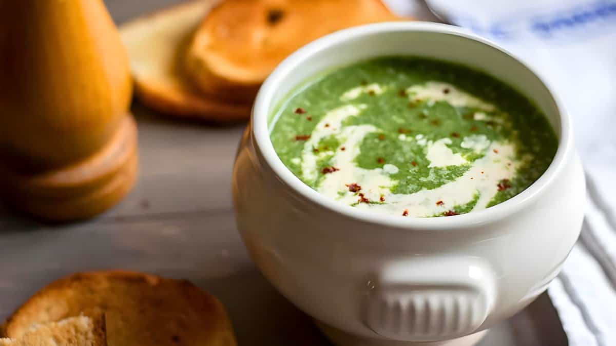 A bowl of creamy soup garnished with herbs and spices, accompanied by slices of bread on a wooden table.
