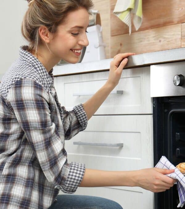 A woman in a plaid shirt smiles while taking out a tray of baked scones from an oven in a modern kitchen.