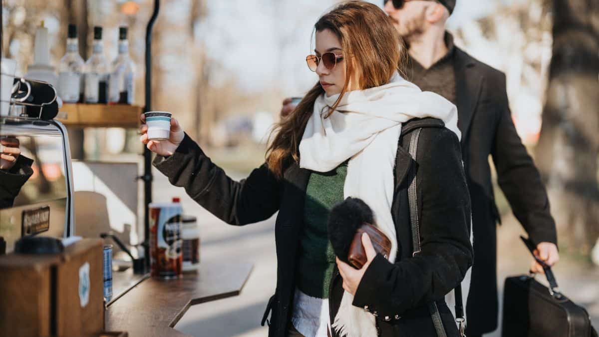 A woman in sunglasses receives a cup of coffee from an outdoor vendor on a sunny day, with a man waiting behind her.