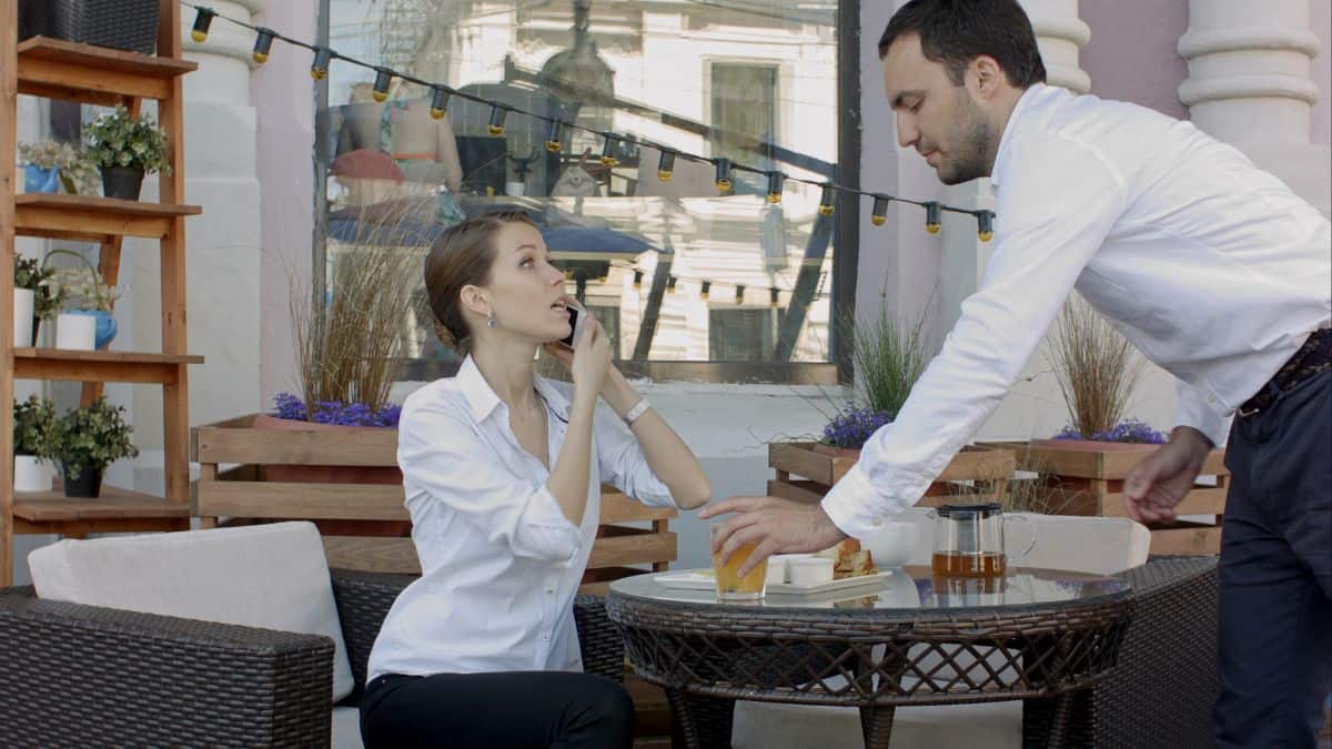 A waiter serving a smiling woman seated at a cafe terrace, with tea and sandwiches on the table.