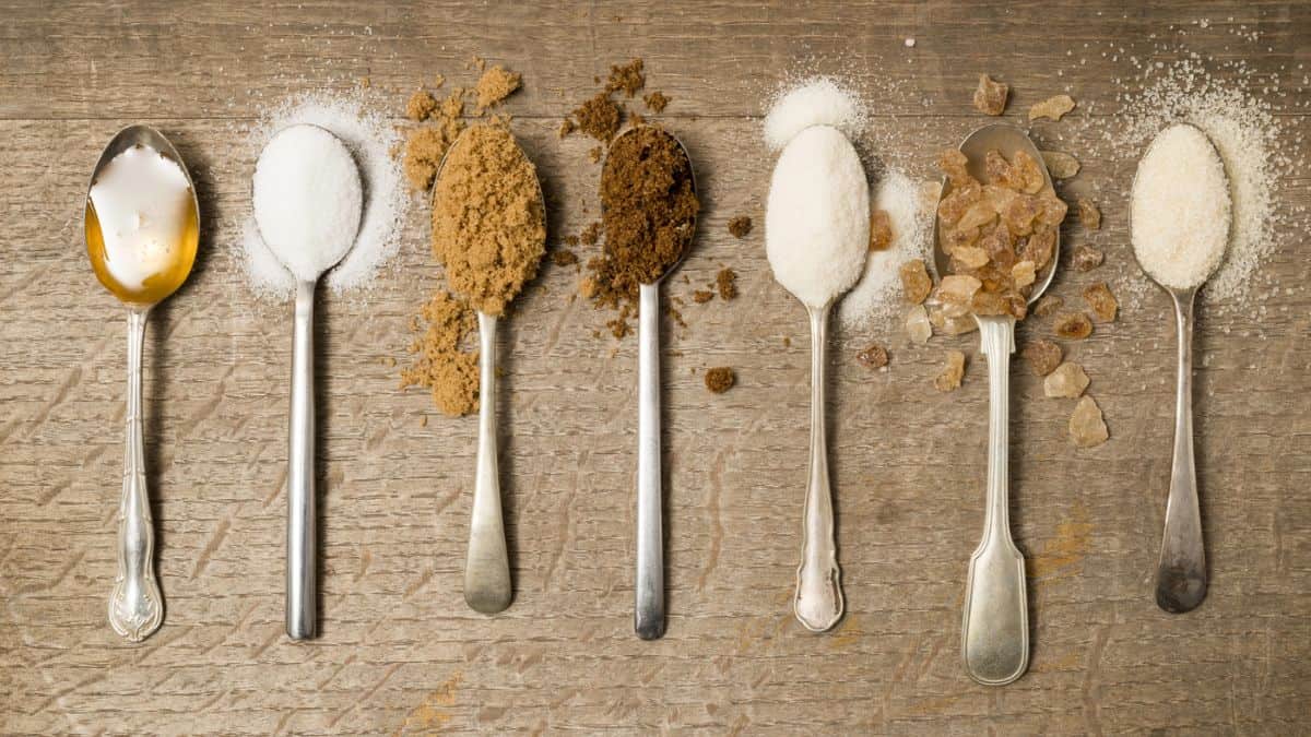 Six spoons on a wooden surface holding various types of sugar, ranging from white and brown granulated sugar to raw sugar crystals.