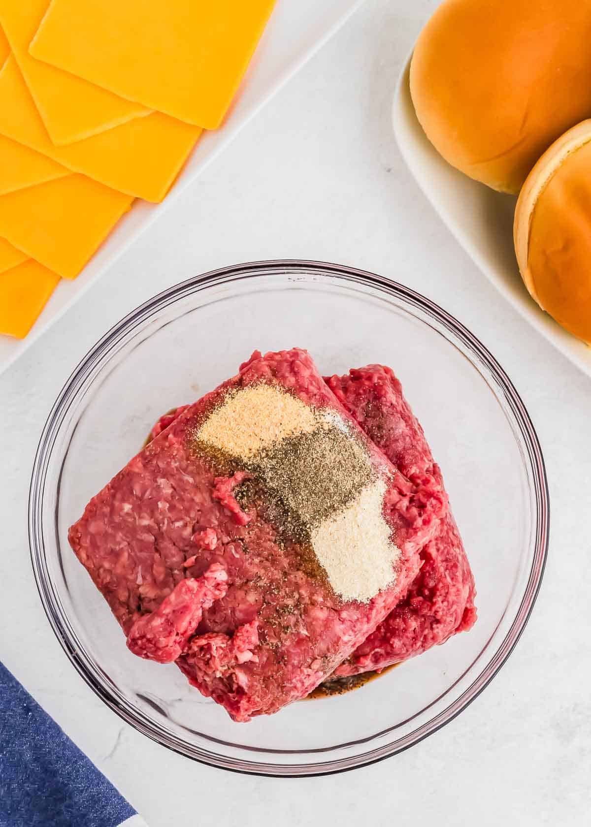 A bowl of raw ground bison seasoned with spices on a kitchen counter, accompanied by slices of cheddar cheese and burger buns.