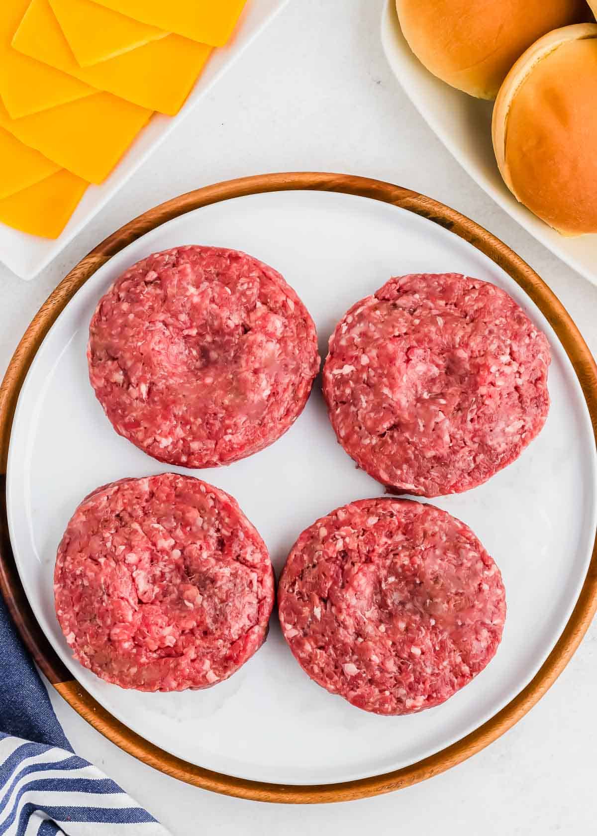 Four raw bison burger patties on a white plate, with slices of cheese and buns arranged around it on a marble countertop.