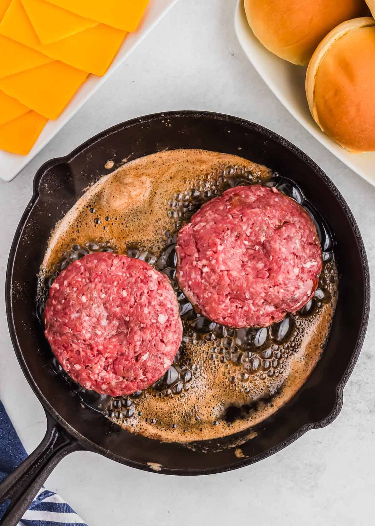 Two raw hamburger patties cooking in a cast iron skillet with bubbling butter, accompanied by sliced bread and cheese on the side.