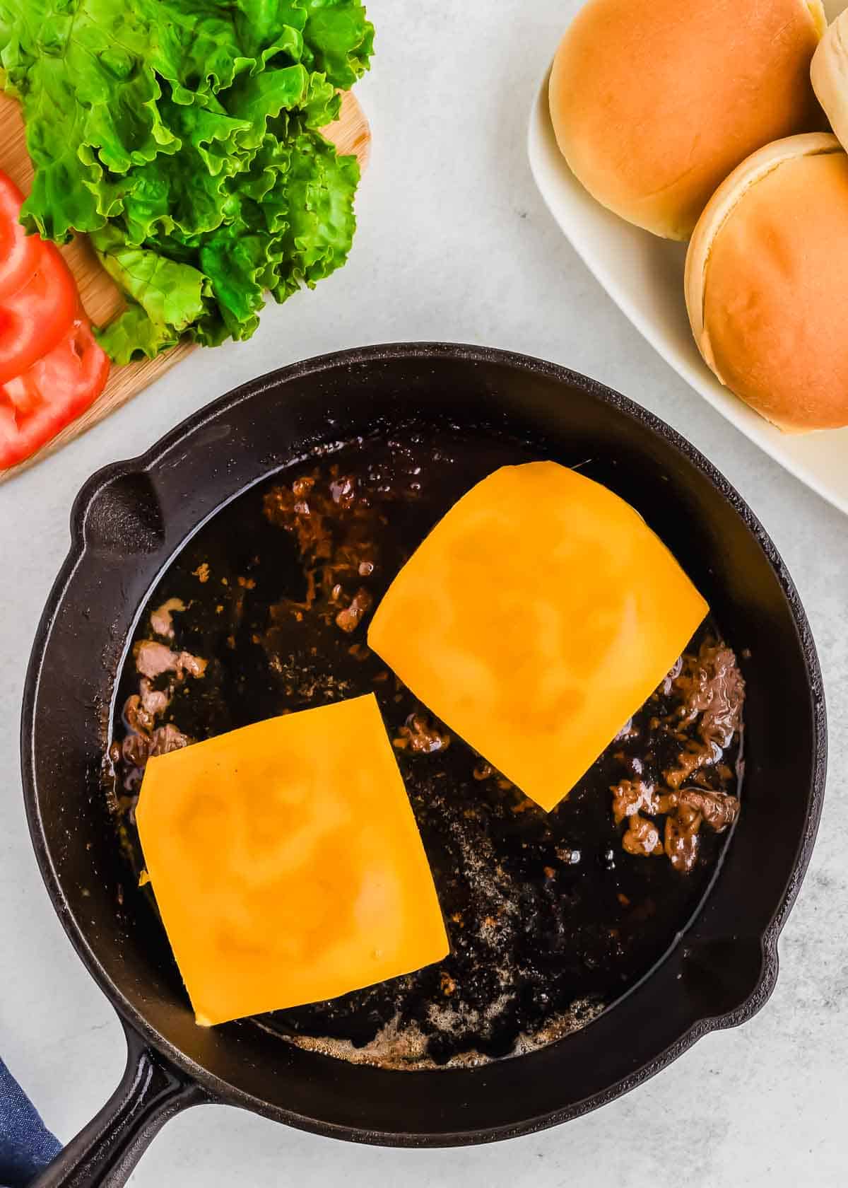 Two cheese-topped burger patties cooking in a skillet, with lettuce, tomatoes, and burger buns on the side.