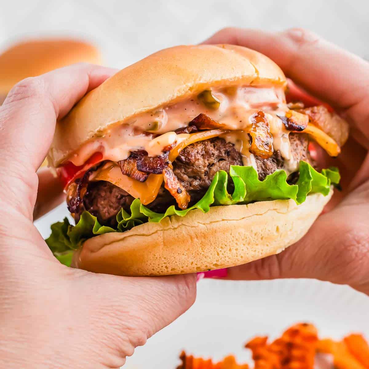 Hands holding a burger with bacon, lettuce, grilled onions, tomatoes, and sauce on a bun.