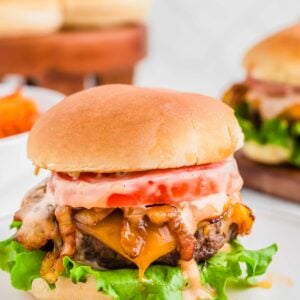 A bison cheeseburger with lettuce, tomato, and sauce on a bun, served on a white plate, with more burgers blurred in the background.