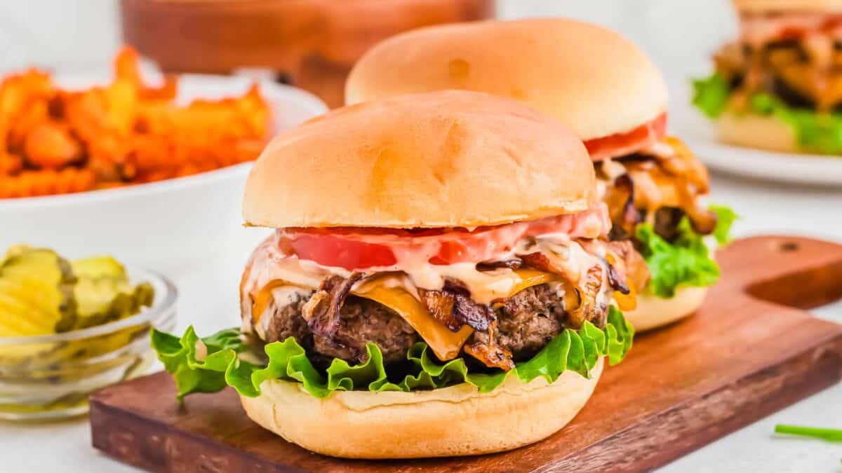 Two bison cheeseburgers with lettuce, tomato, and sauce on a wooden board, accompanied by sides of pickles and sweet potato fries.