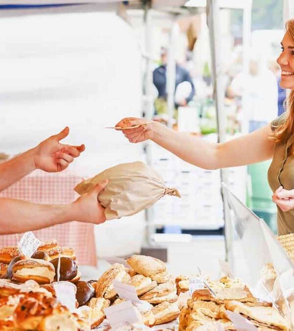 A woman buys bread from a male vendor at an outdoor market stall.