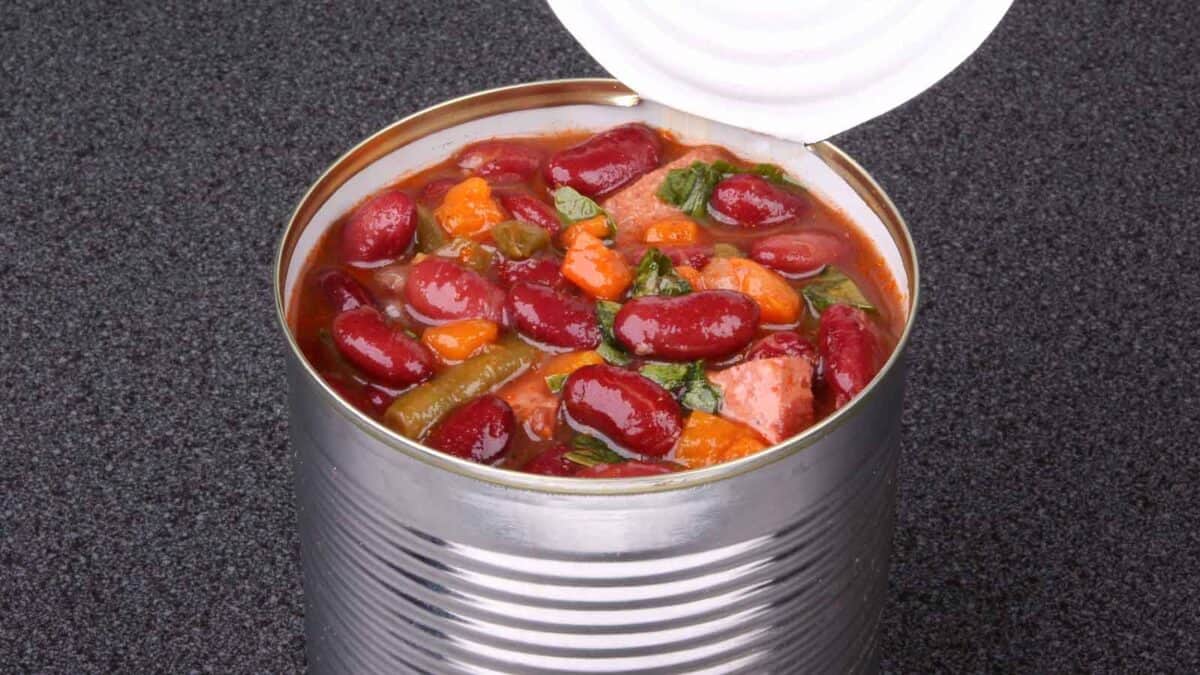 An opened can of mixed vegetables and red kidney beans in tomato sauce on a dark countertop.