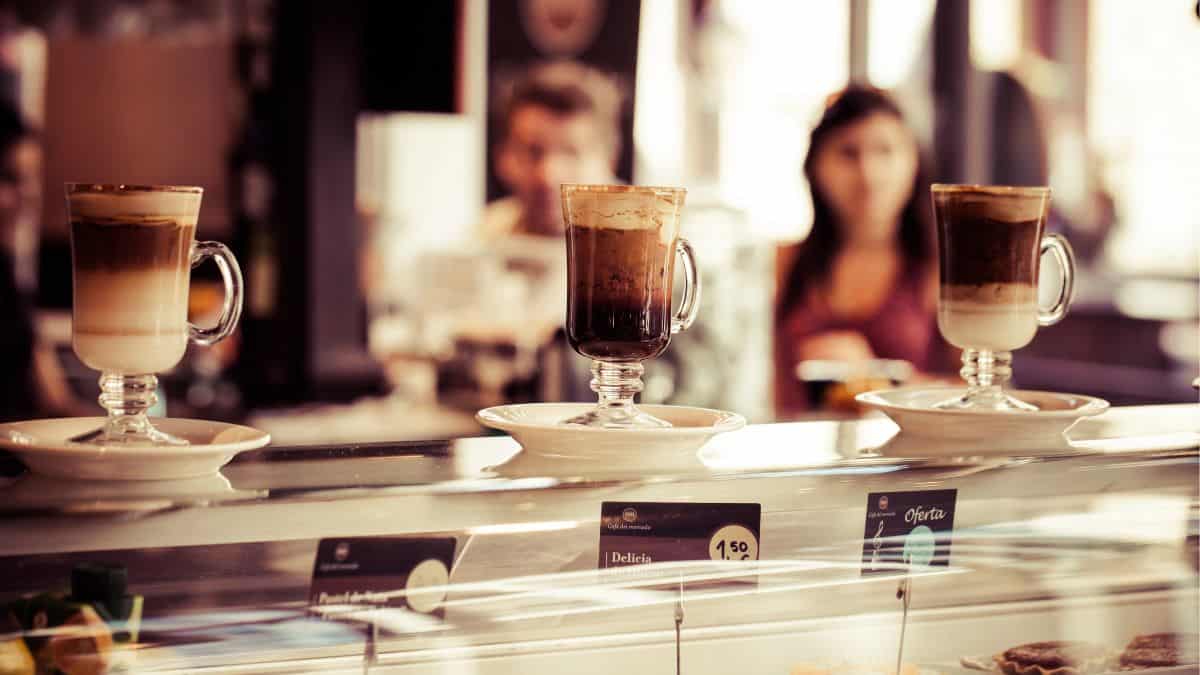 Three cups of coffee with layers of cream displayed on a glass counter in a cafe, with blurred patrons in the background.