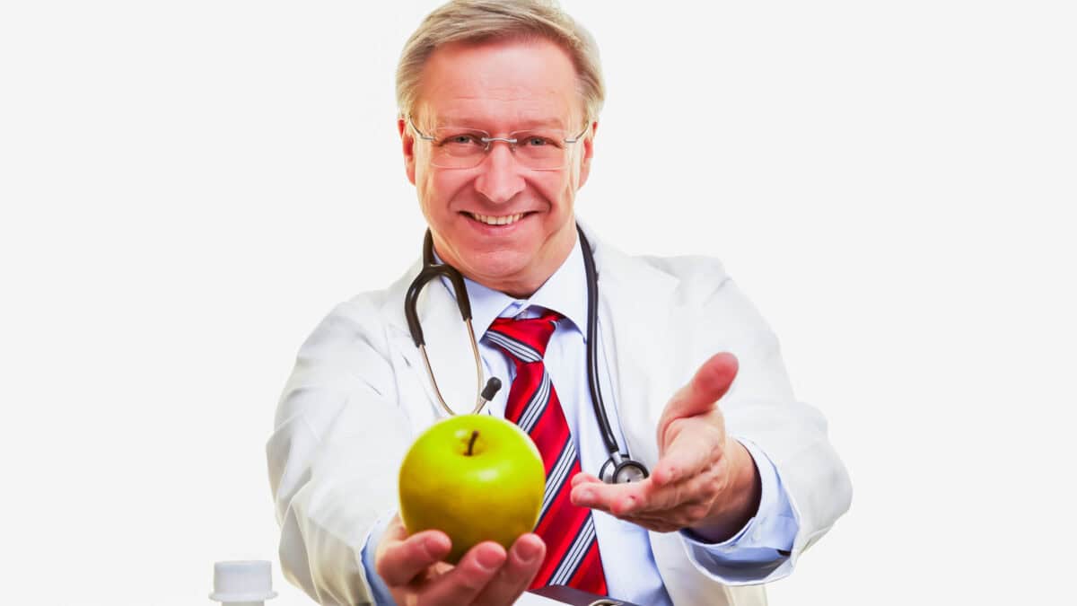 A smiling male doctor wearing a lab coat and stethoscope, offering a green apple with one hand extended forward.