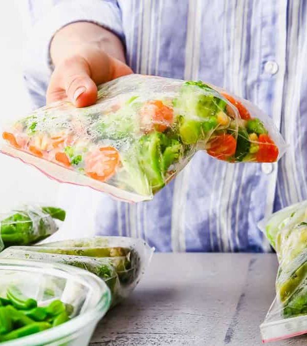 Person places mixed vegetables into a plastic freezer bag for storage, with other bags and vegetables visible on the table.